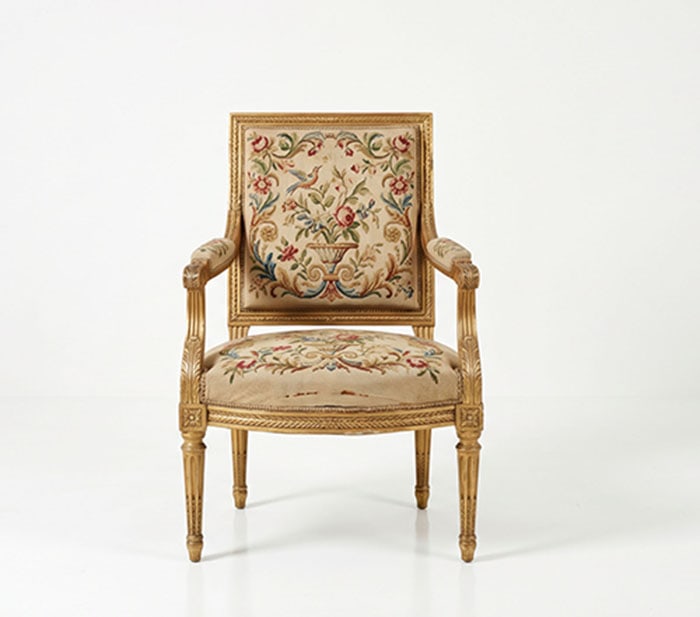 Giltwood Armchair: Armchair featuring a brass finish and woven upholstery on the backrest, seat, and arms. The fabric displays a multi-colored floral and bird motif. The carved frame includes fluted legs and decorative