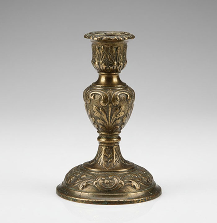 Bronze Candlestick: Brass candlestick has a round base and a decorated stem with leaf and scroll motifs. The top contains a single socket for holding a candle. The surface shows an aged finish with some patina near the