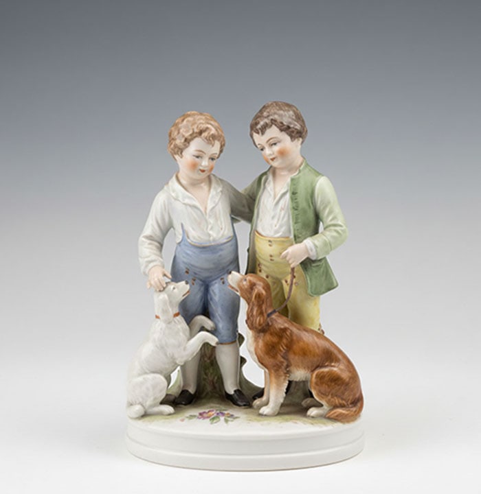 Figural Group of Two Boys: Ceramic figurine depicting two boys standing with a white dog and a brown dog on a circular base. The figures are painted in blue, green, and yellow. Small floral details are painted on the white