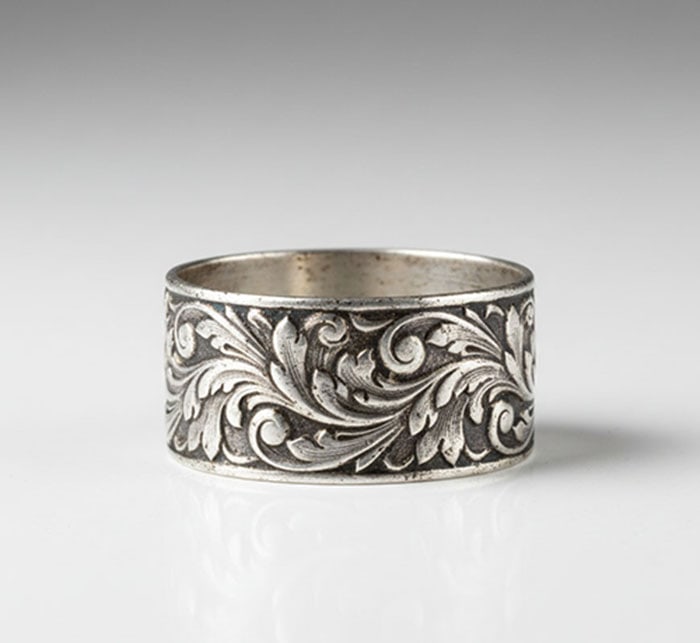 Napkin Holder: White metal napkin ring decorated with an engraved leaf and scroll motif. The object has a circular band shape with a dark recessed background in the pattern. The inner surface shows signs of wear
