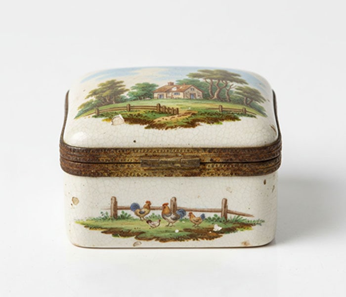 English Pastoral Scenes Ceramic Box: Rectangular ceramic box with a hinged lid and weathered brass metal trim. The exterior surfaces are decorated with a crackled white glaze and painted scenes of a countryside house and chickens. L: 14