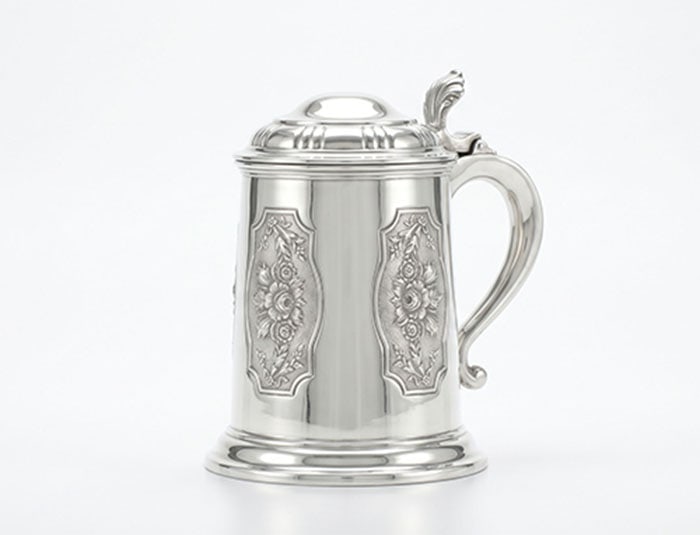 A Fine Lidded White Metal Tankard: White metal tankard features embossed floral panels on its body. It includes a hinged lid with a thumb lever and a large curved handle. The base is flared and circular.