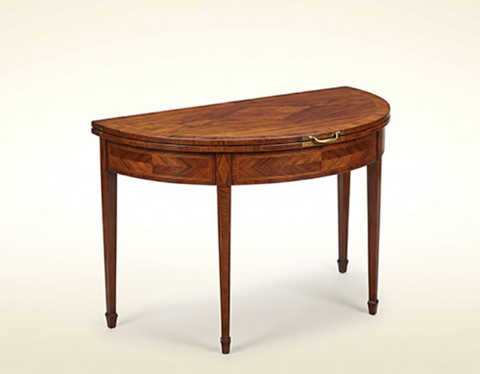 Mahogany Demilune Table: Wooden demi-lune table featuring a folding top and four tapered legs. Inlaid wood patterns decorate the apron and top surface. Brass latch is mounted on the front edge. H: 70 CM