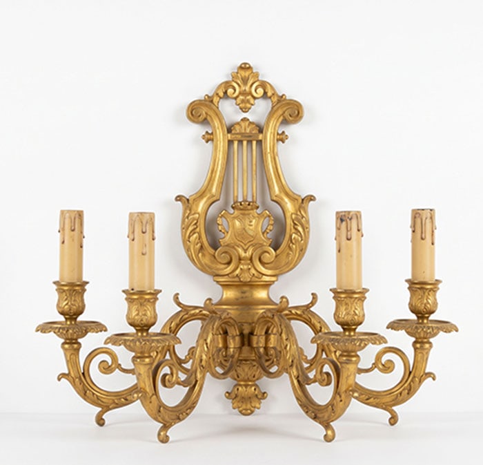 A Gilt-Bronze Lyre-Form Sconce: Wall sconce made of brass featuring a central lyre design and four curved arms. Each arm holds a cream-colored candle sleeve with faux wax drips. The piece has ornate scrolls and leaf motifs