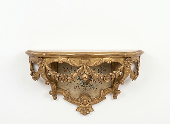 Giltwood Console: Wall shelf finished in brass color featuring carved foliate motifs and floral patterns. A central panel contains a small painted flower arrangement. The top is flat and shaped to sit against a wall.