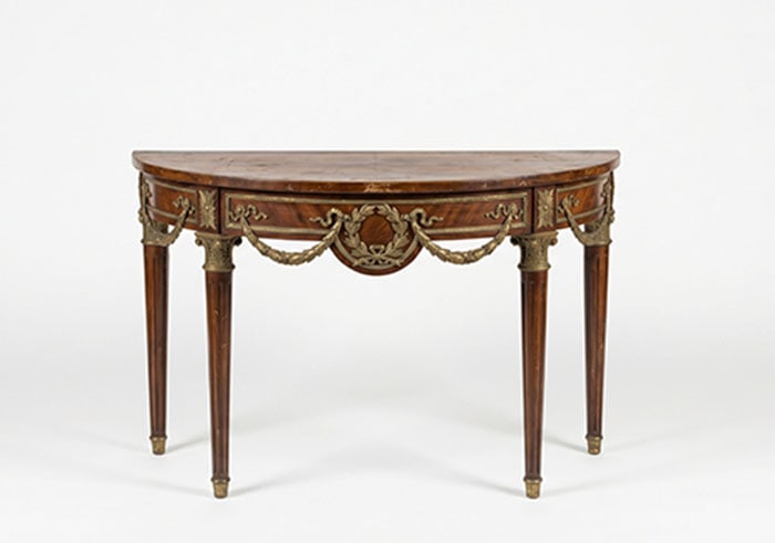 Demilune Console: Semicircular wooden table featuring brass ornamental swags and wreaths along the apron. Wood surfaces exhibit visible wear and scratching, particularly on the top. Four tapered legs support the