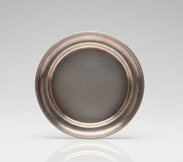 A Small Circular Pewter Dish: White metal plate with a flat center and a raised, grooved border. The surface exhibits a dull finish with light surface marks. D: 18 CM