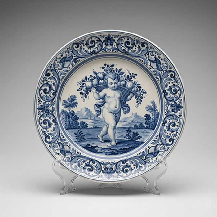 Delftware Blue and White Plate: Circular ceramic plate featuring a blue illustration of a child carrying a basket of fruit in a landscape setting. The rim is decorated with a repeating pattern of scrolls and floral motifs. D: 15 CM