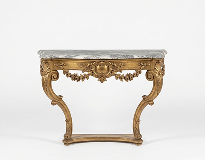 Giltwood Console: Console table featuring a marble top with grey veining and an ornate brass base. The base has two curved legs and is decorated with leaf and flower motifs. H: 71 CM