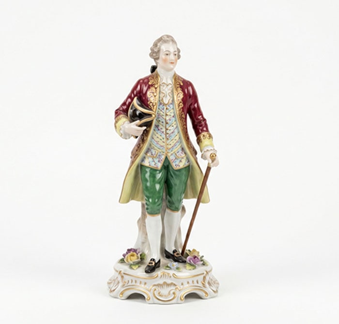 Porcelain Figure: Ceramic figurine of a man dressed in a red coat, patterned vest, and green breeches holding a walking stick. The figure stands on a white base decorated with small flowers and brass-colored accents.