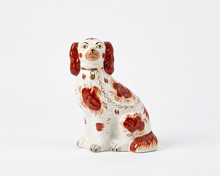 Ceramic Spaniel Figurine: Ceramic spaniel figurine featuring white glaze with reddish-brown patches and long drooping ears. Brass collar and chain are painted around the neck and draped across the side. H: 14 cm