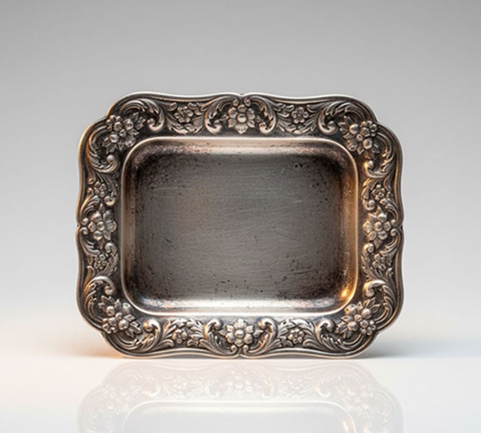 White Metal Dish: White metal dish featuring a raised border with embossed floral and leaf motifs. Central recessed area has a matte finish and shows signs of surface wear. Edges are shaped with a scalloped profile