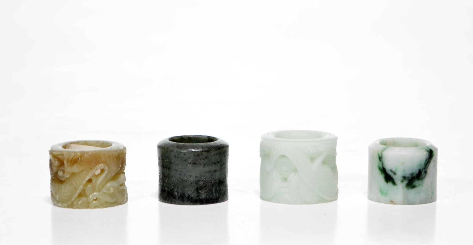 Chinese Jade Archer's Rings: Collection of four archer's rings expertly fashioned from diverse varieties of jade, ranging from pale celadon and mottled emerald to a deep, dark spinach hue. Two examples are intricately carved in r