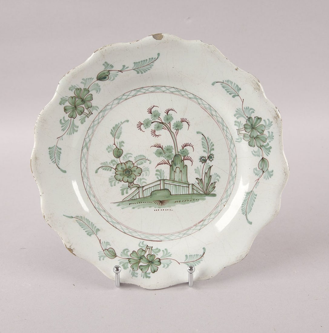 Porcelain Plate: With a charming lobed rim and soft tin-glaze, the plate is centered with an Chinoiserie landscape scene featuring stylized trees, rocks, and foliage rendered in soft green and puce enamels. 19 cm
