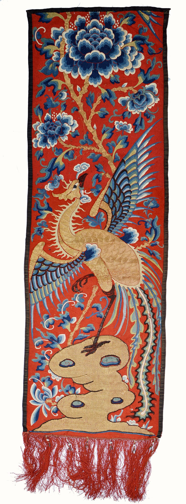 Chinese Embroidered Phoenix Hanging: A rectangular red-ground textile panel worked in polychrome and gold thread with a phoenix perched on pierced rocks amid flowering peony sprays and scrolling foliage. Bordered in black and finished