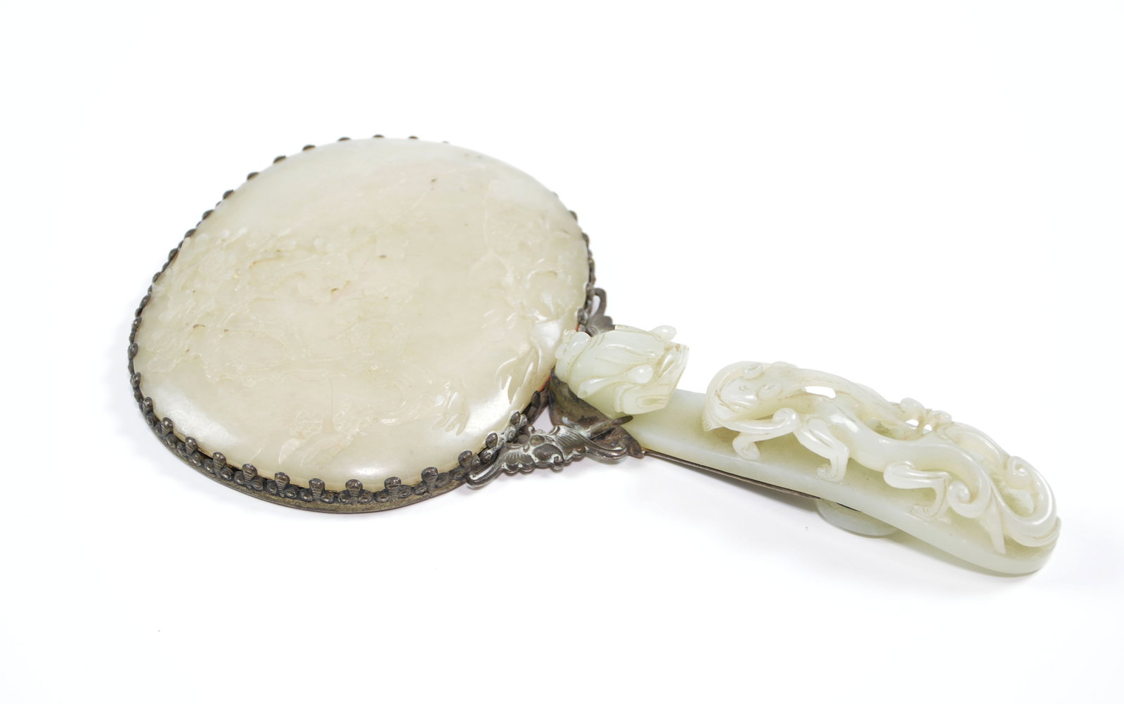 Chinese Jade-Mounted Hand Mirror: The circular pale celadon jade mirror-back is carved in low relief with scrolling floral sprays and set within a darkened metal mount with a beaded rim. The similarly hued jade handle is carved and