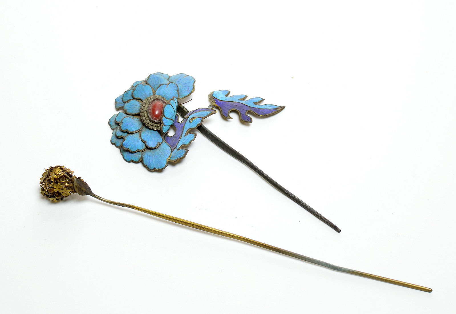 Chinese Floral Hairpins: Comprising two hairpins. One is decorated with a layered blue flower centred by a red cabochon in a scalloped mount, with a blue and purple enamelled leafy stem on a dark metal pin. The other has a gi