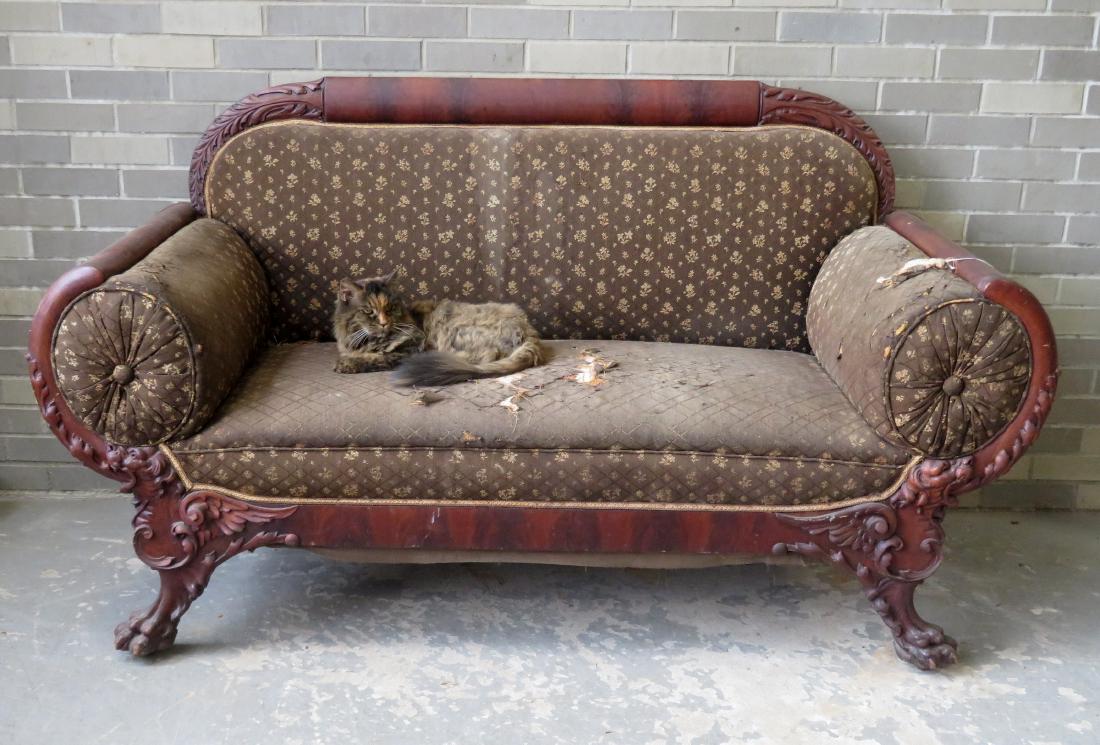 An American Empire upholstered settee having a carved: An American Empire upholstered settee having a carved wooden mahogany frame with rounded sides to accommodate the 2 fitted pillows. The the sides of the back crest with acanthus carvings, and the heav