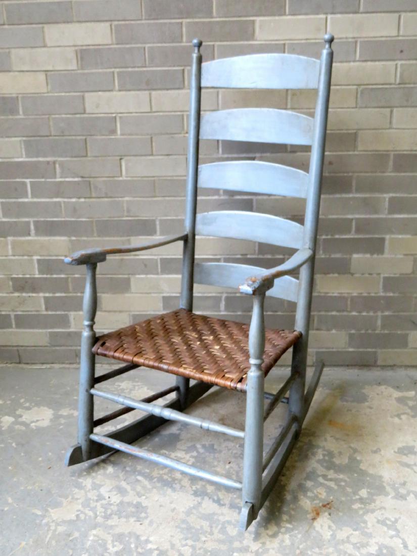A country ladder back armchair rocker having 5 curved: A country ladder back armchair rocker having 5 curved slats, and shaped arms mortised through the front arm supports. Later blue paint and newer woven cane seat, very good condition. 18th century.