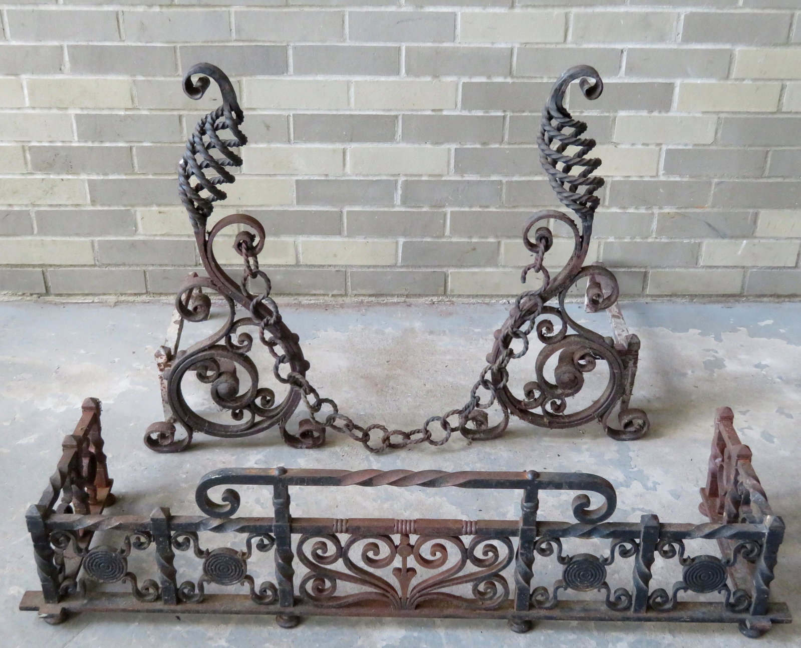 A pair of Arts & Crafts wrought iron Roycroft andirons: A pair of Arts & Crafts wrought iron Roycroft andirons and near matching wrought iron fireplace fender. The andirons in old black paint with the typical Roycroft scrolls and the original chain across 