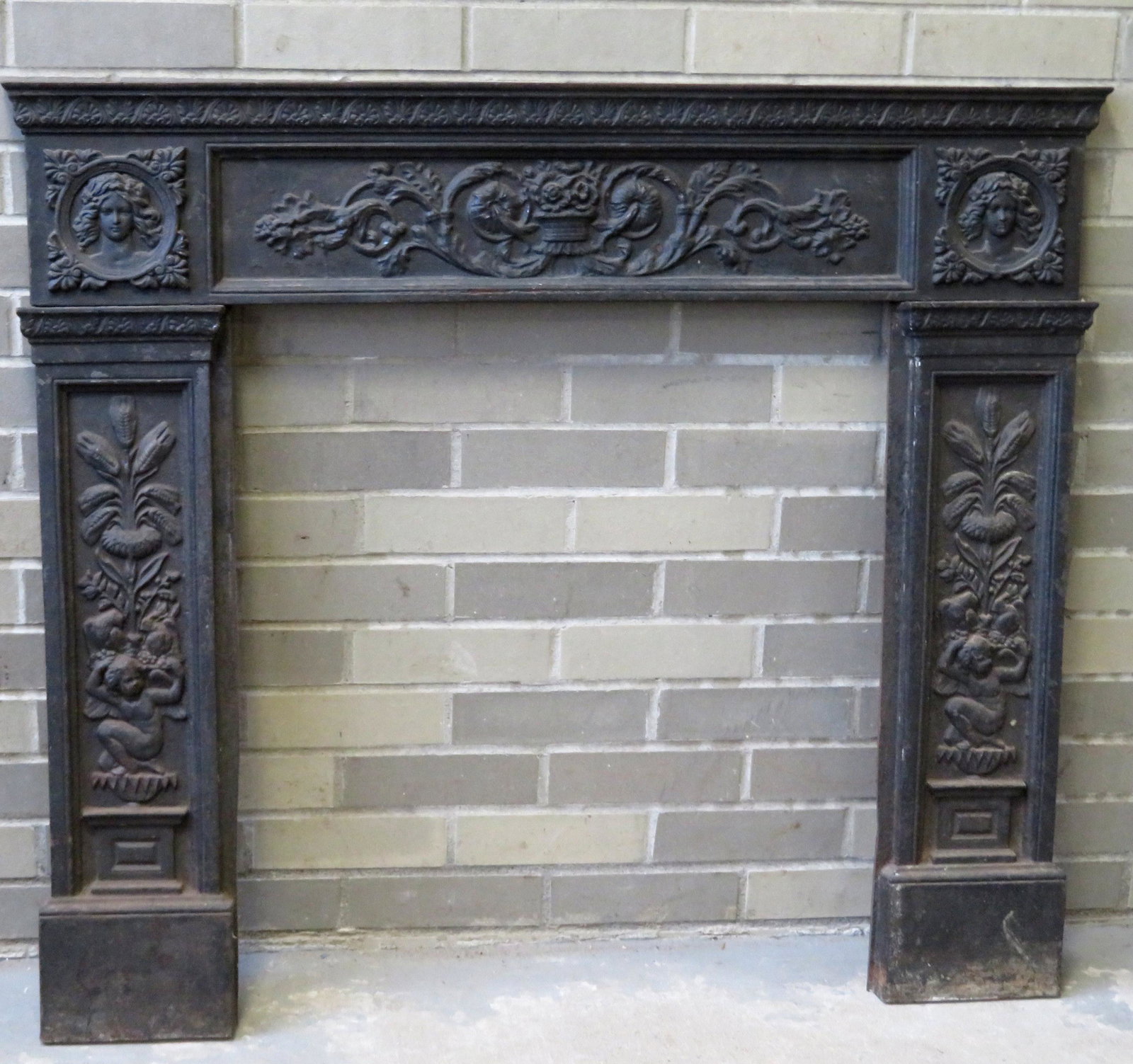 A cast iron fireplace surround having a frieze panel: A cast iron fireplace surround having a frieze panel decorated with a basket of flowers within extending scrolled vines, flowers and leaves adjacent to a bust of a lady on each end over pilasters deco