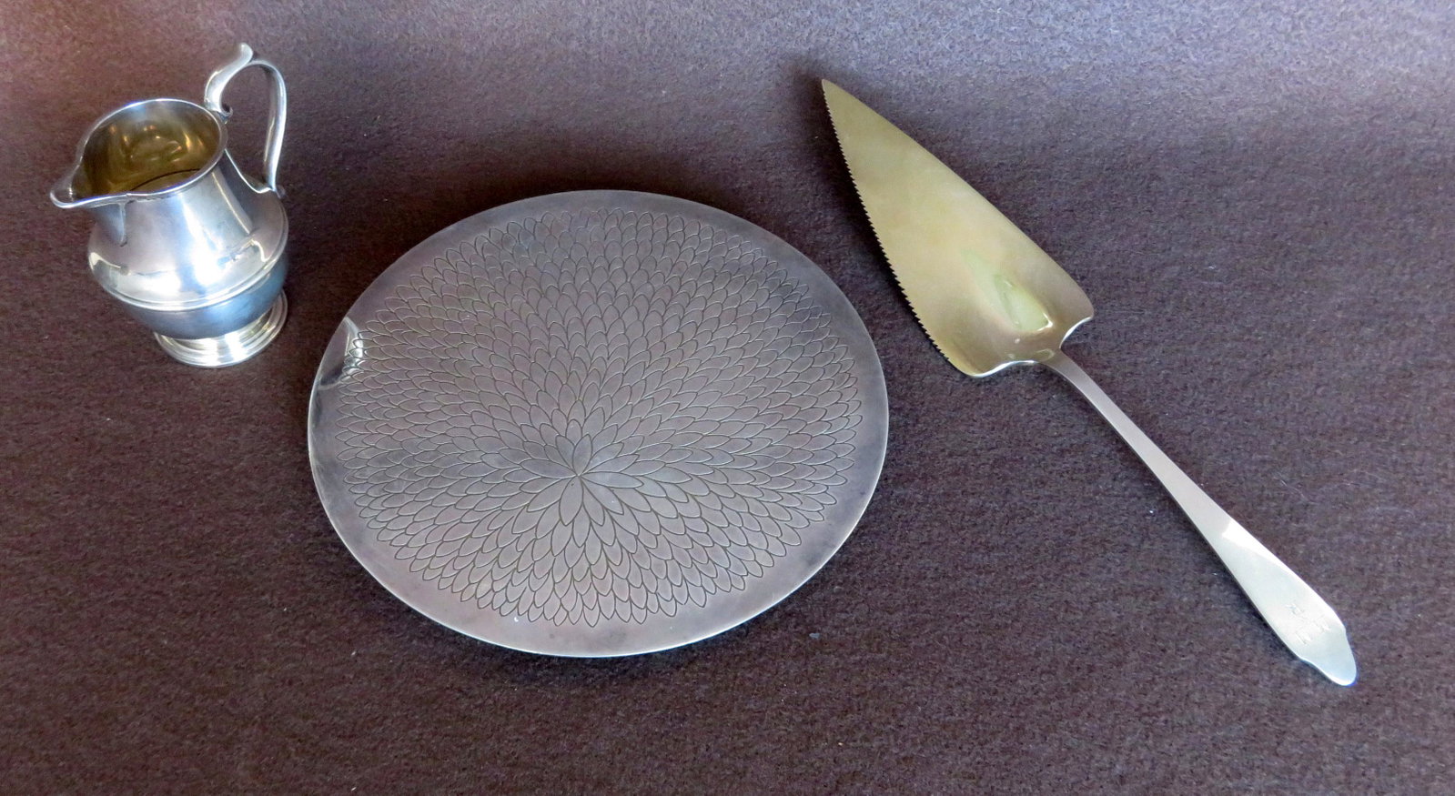 Three pieces of Tiffany & Co. sterling silver including: Three pieces of Tiffany & Co. sterling silver including a creamer - 4"H, a cake knife with gold wash - 11 1/4"L, and a circular footed trivet decorated with a large stylized flower - 7 3/4"D. All in v