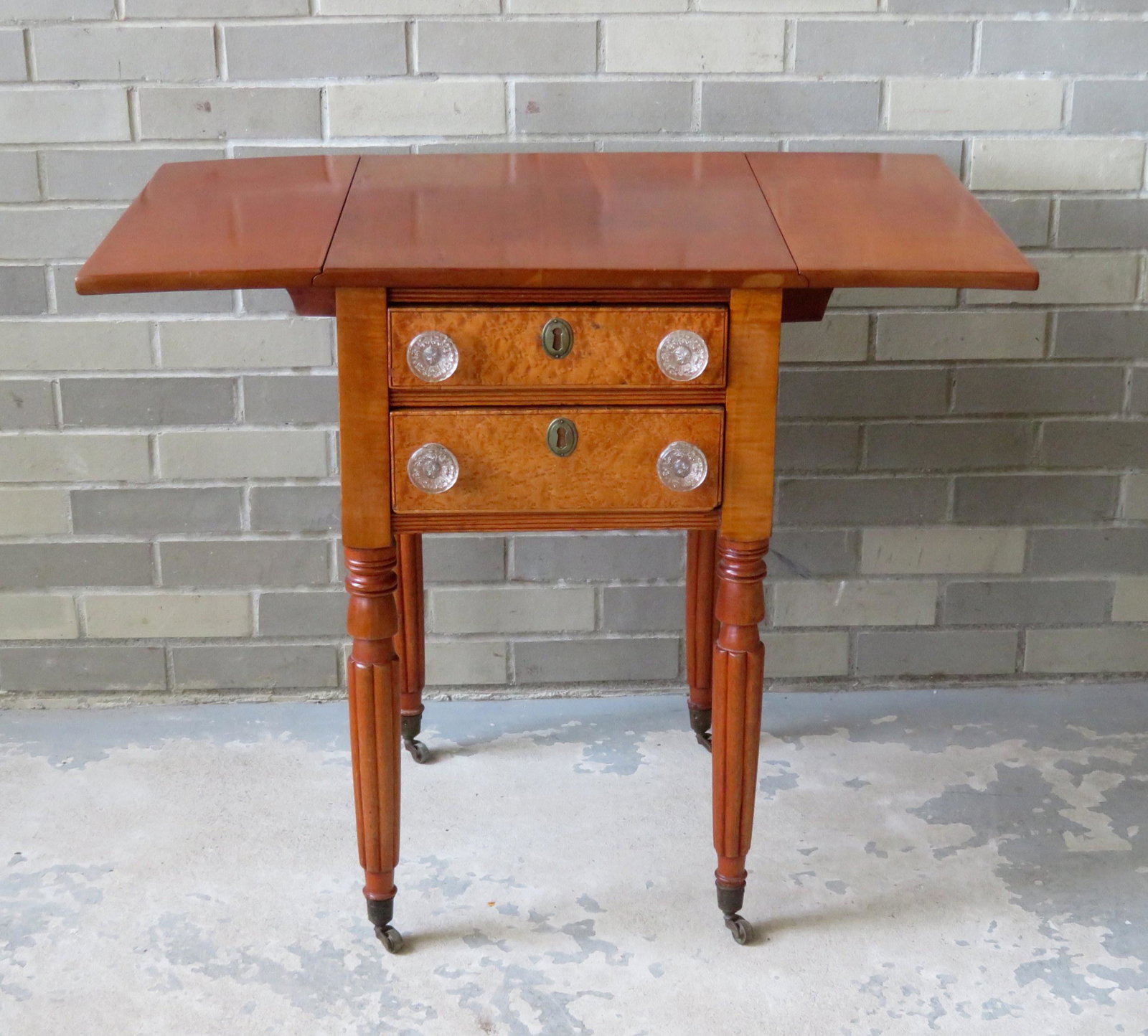 Sheraton 2 drawer drop leaf sewing stand having a: Sheraton 2 drawer drop leaf sewing stand having a cherry top, 2 veneered birdseye maple drawer fronts with original glass pulls, and a maple frame. The legs are tapered and reeded and lead to the bras