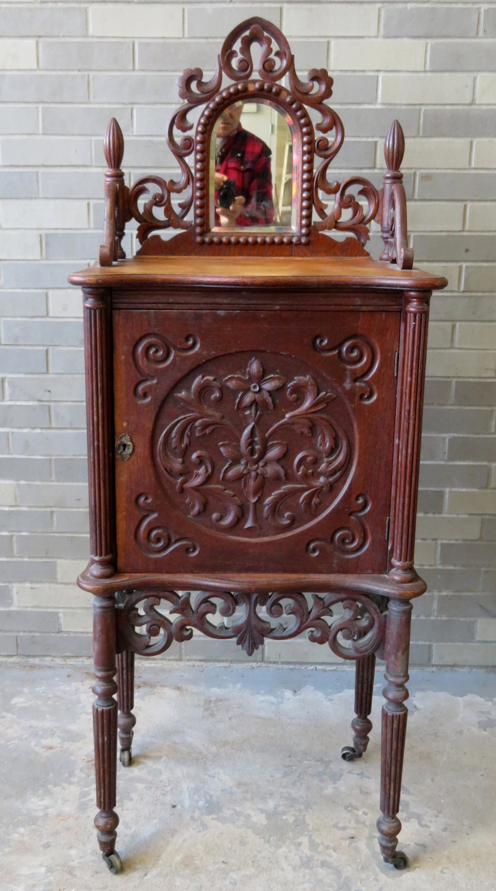 An ornate oak sheet music cabinet having a backsplash: An ornate oak sheet music cabinet having a backsplash with pierced scrollwork and a beveled mirror attached to a serpentine shaped top (and front) over a large single door decorated with a carved flow