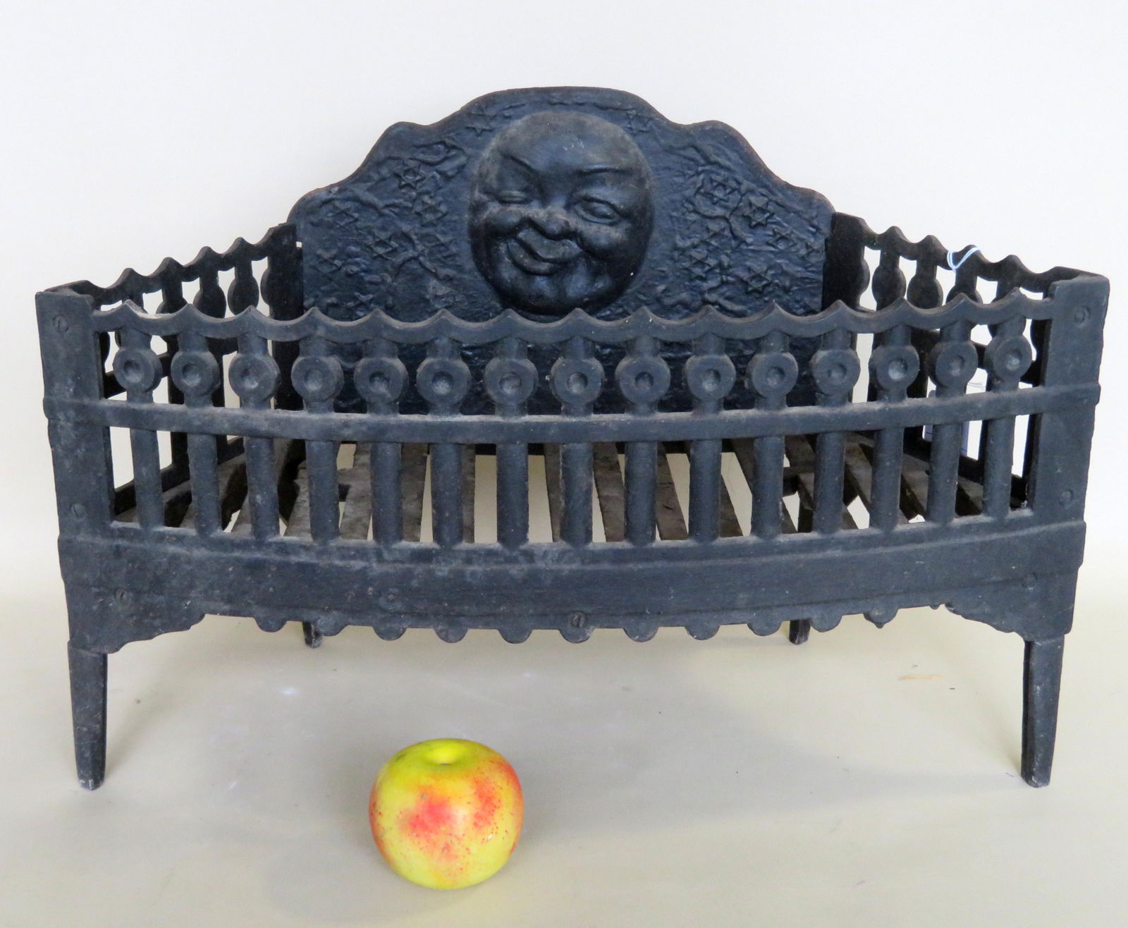A cast iron fireplace grate having a decorated: A cast iron fireplace grate having a decorated fence-like border on 3 sides and a man-in-the-moon embossed face surrounded by stars on the backsplash. 22"W x 11"D x 14 5/8"H. Very good condition. Late