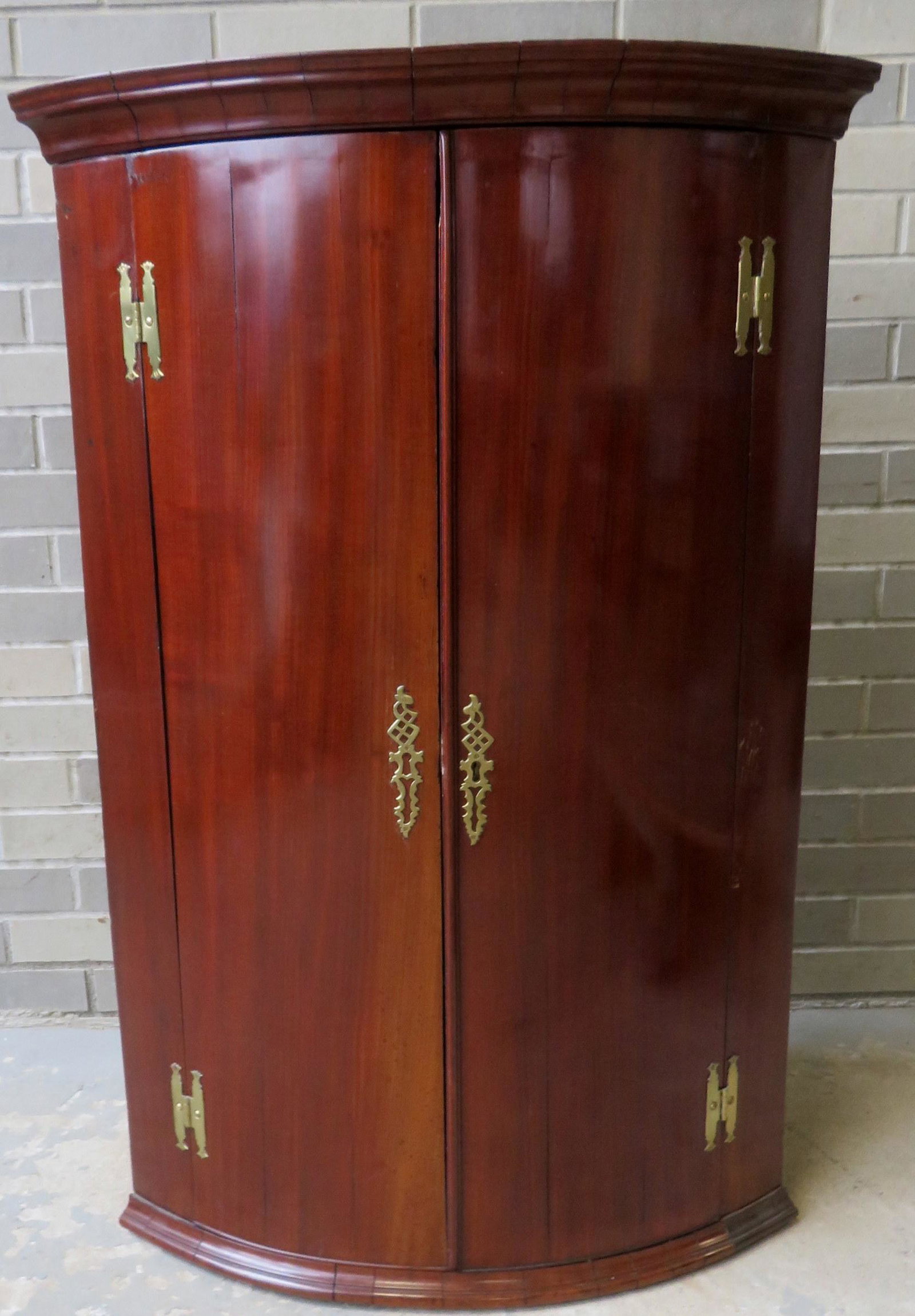 Hanging mahogany double door corner cabinet with bow: Hanging mahogany double door corner cabinet with bow front and H-brass hinges that appear to be original - 3 interior shelves - fancy brass escutcheons. 26"W x 18 1/2"D x 40"H. Two small areas of vene
