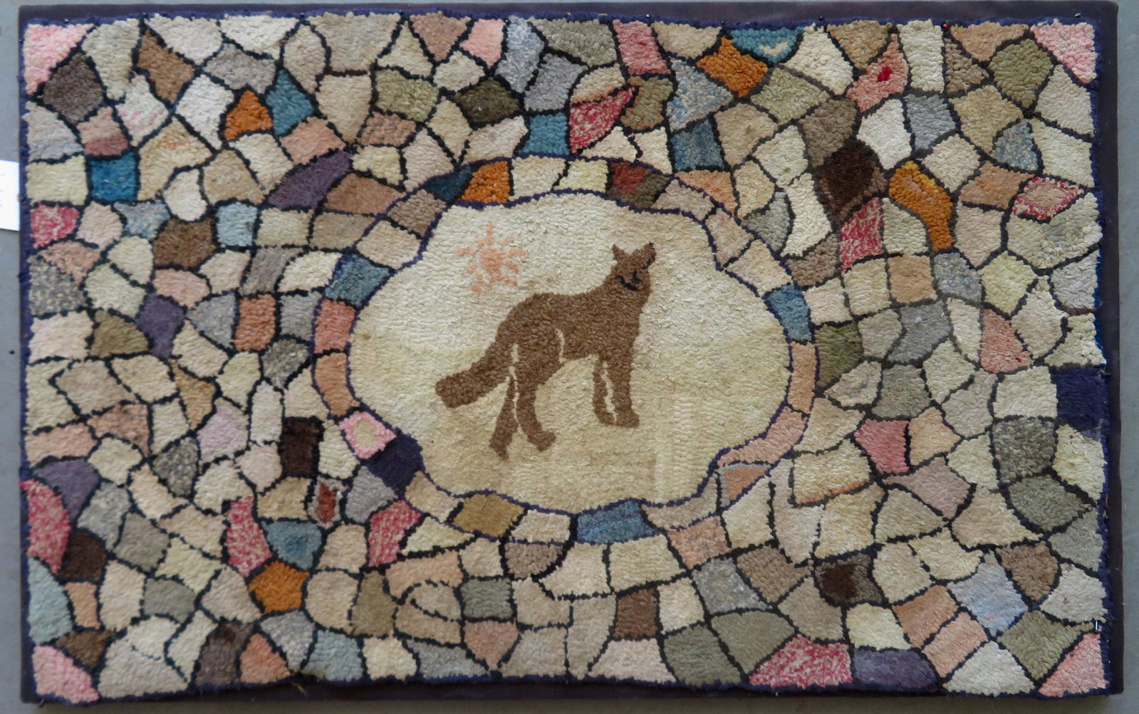 Hooked rug having a central decoration of a howling dog: Hooked rug having a central decoration of a howling dog or wolf below sun, surrounded by small colorful irregular patterns, some geometric. Professionally mounted on frame. Slight darkening of wool, b
