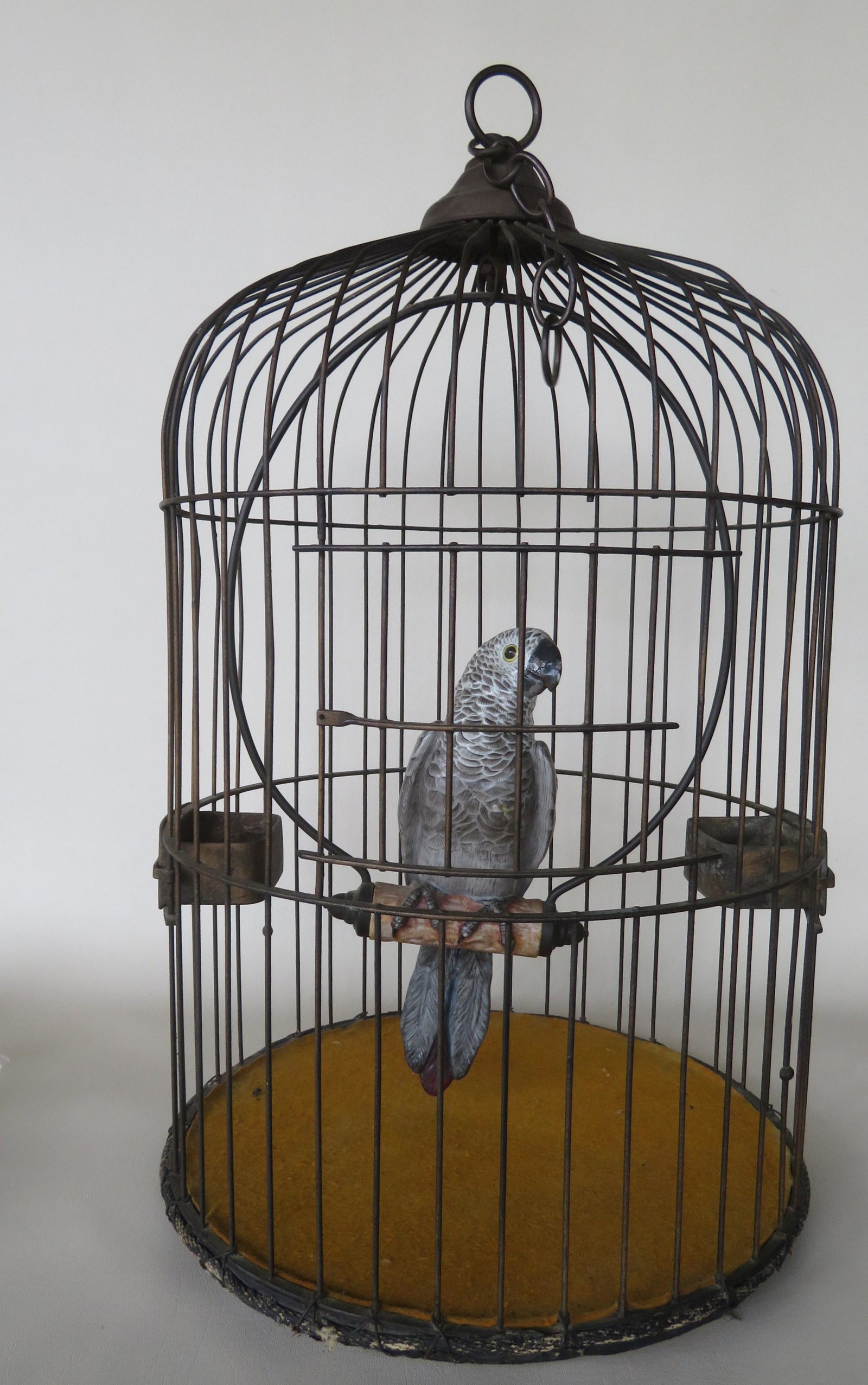 An unusual iron wire bird cage having a porcelain: An unusual iron wire bird cage having a porcelain parrot mounted on perch, and 2 feeds attached to the interior of the sides. The parrot is all hand painted, and likely German, and the cage in old gol