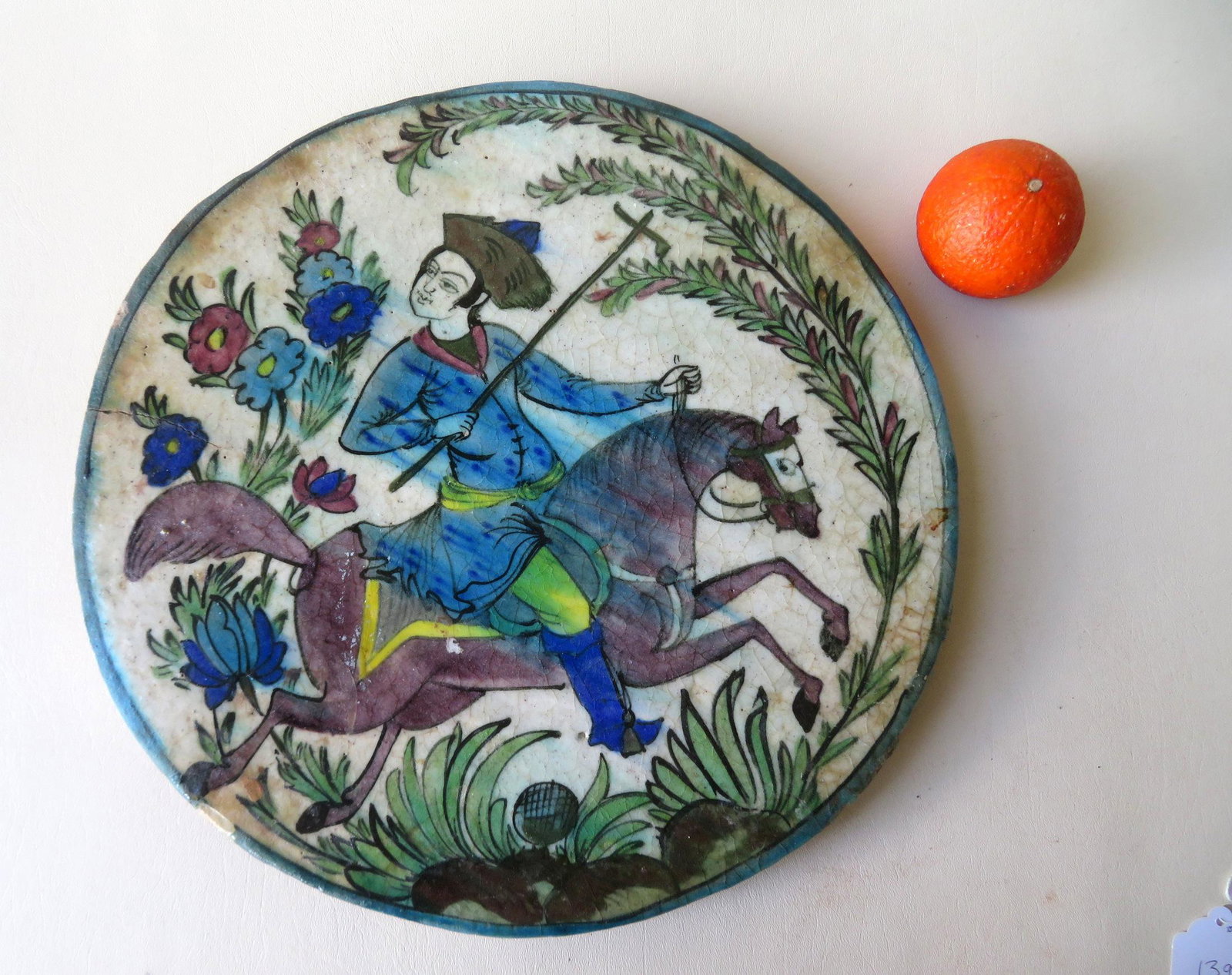 Ceramic Persian plaque decorated with a man on: Ceramic Persian plaque decorated with a man on horseback holding a weapon surrounded by leaves and flowers. 16 7/8" dia. X 3/4" thick. A wood panel has been attached to the back during the 20th centur