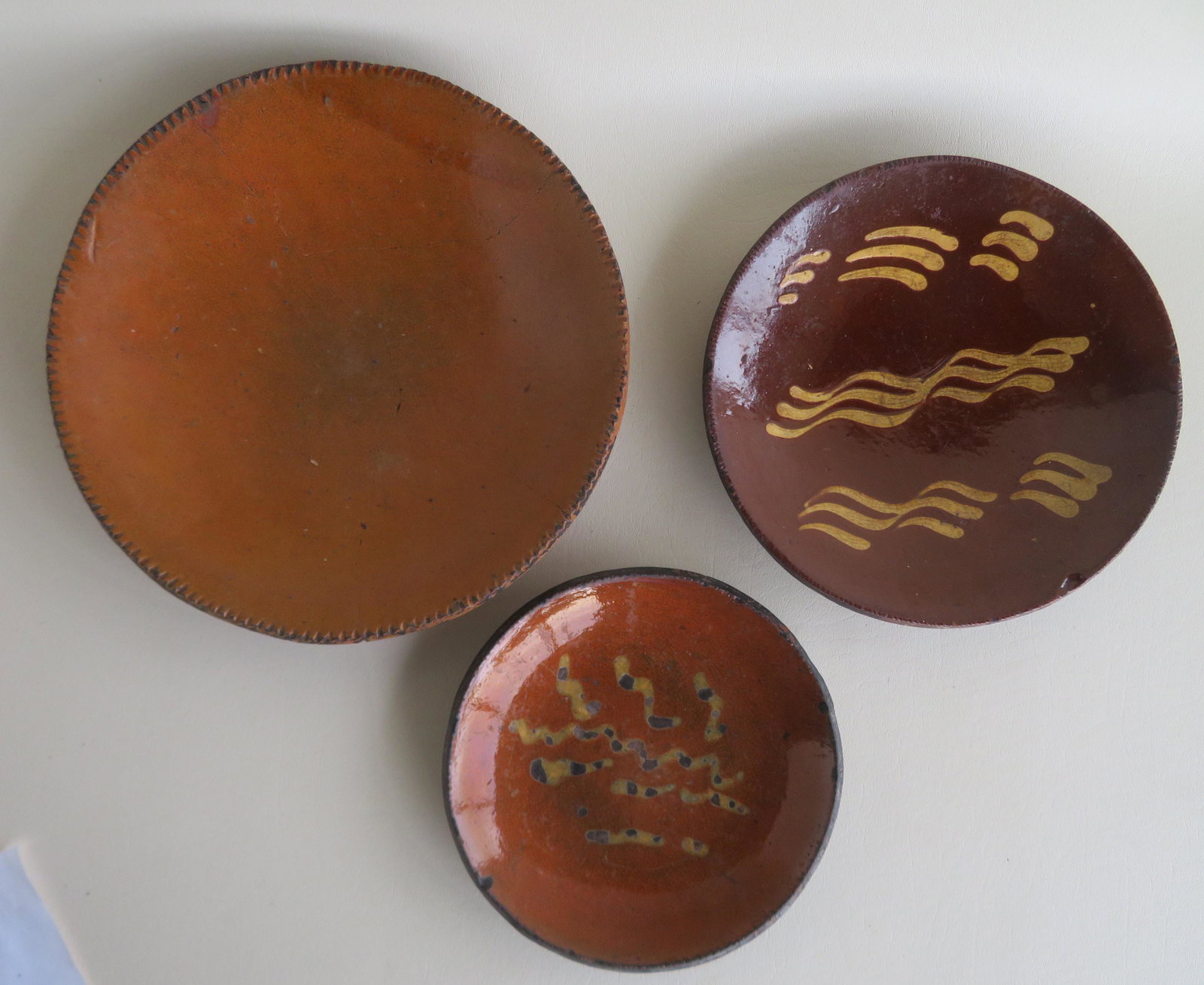 Three redware plates including 2 with yellow slip: Three redware plates including 2 with yellow slip decoration, all 19th century. The smallest, with worn squiggle designs, has minor edge rim chips, and overall good condition - 7" dia.; the mid-sized