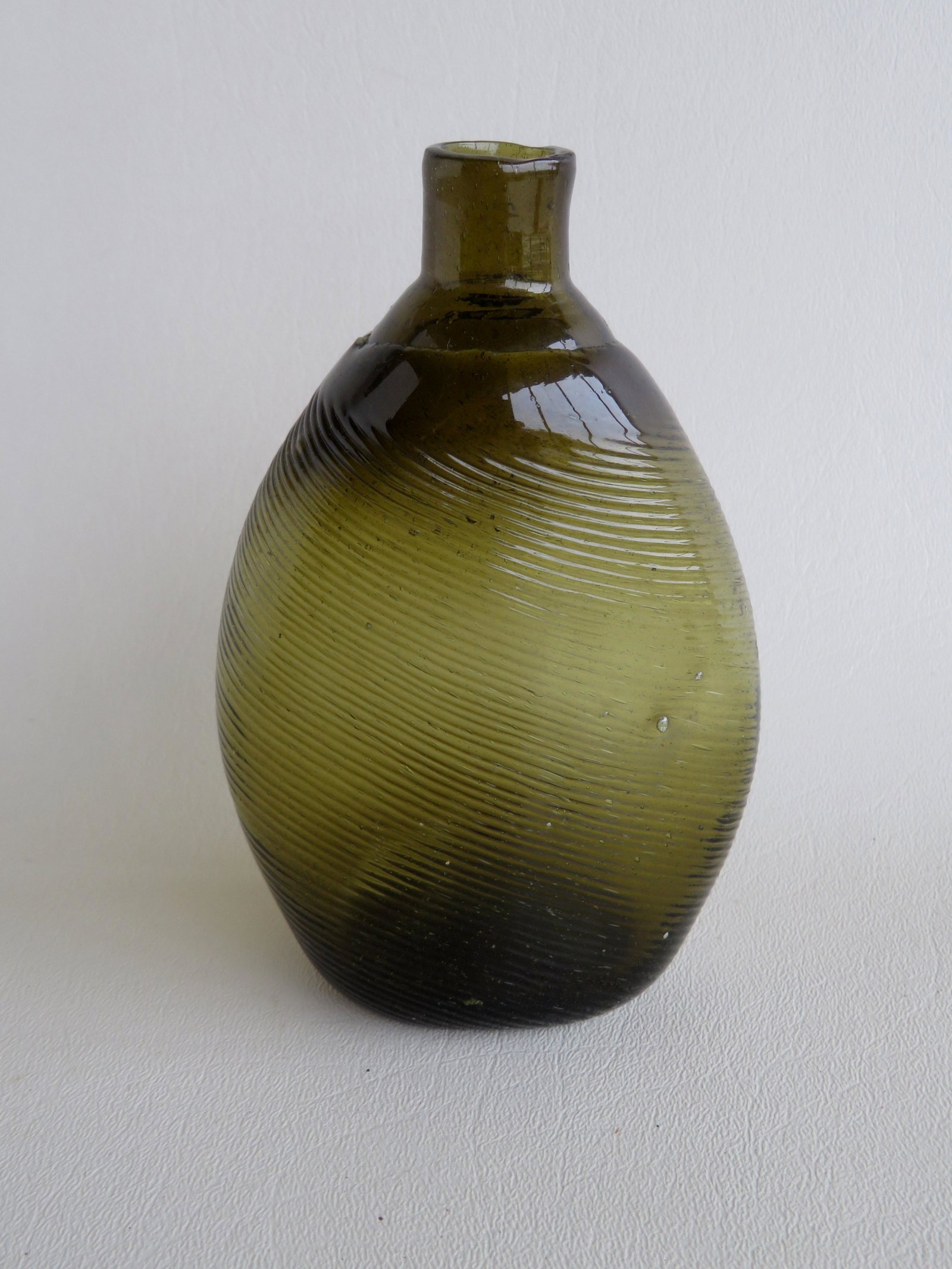 A pitkin style dark olive green flask, swirled to the: A pitkin style dark olive green flask, swirled to the left, irregular ridge around shoulder, pushed up pontil scar on base, probably New England 1790-1830. Very good condition with no damage or repair