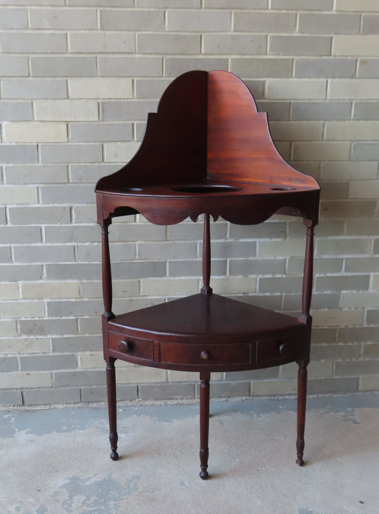 A Sheraton corner washstand with shaped dovetailed: A Sheraton corner washstand with shaped dovetailed high-back backsplash and shaped apron over lower shelf with a central drawer flanked by 2 false drawers on turned legs and ball feet. Circa 1820. One