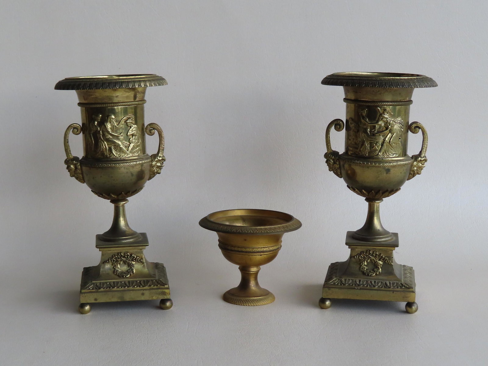 Three classical metal objects including: Two small: Three classical metal objects including: Two small ornate brass urns with devil face handles and having applied classical Roman scenes across the front - each urn mounted on a shaped plinth with acant