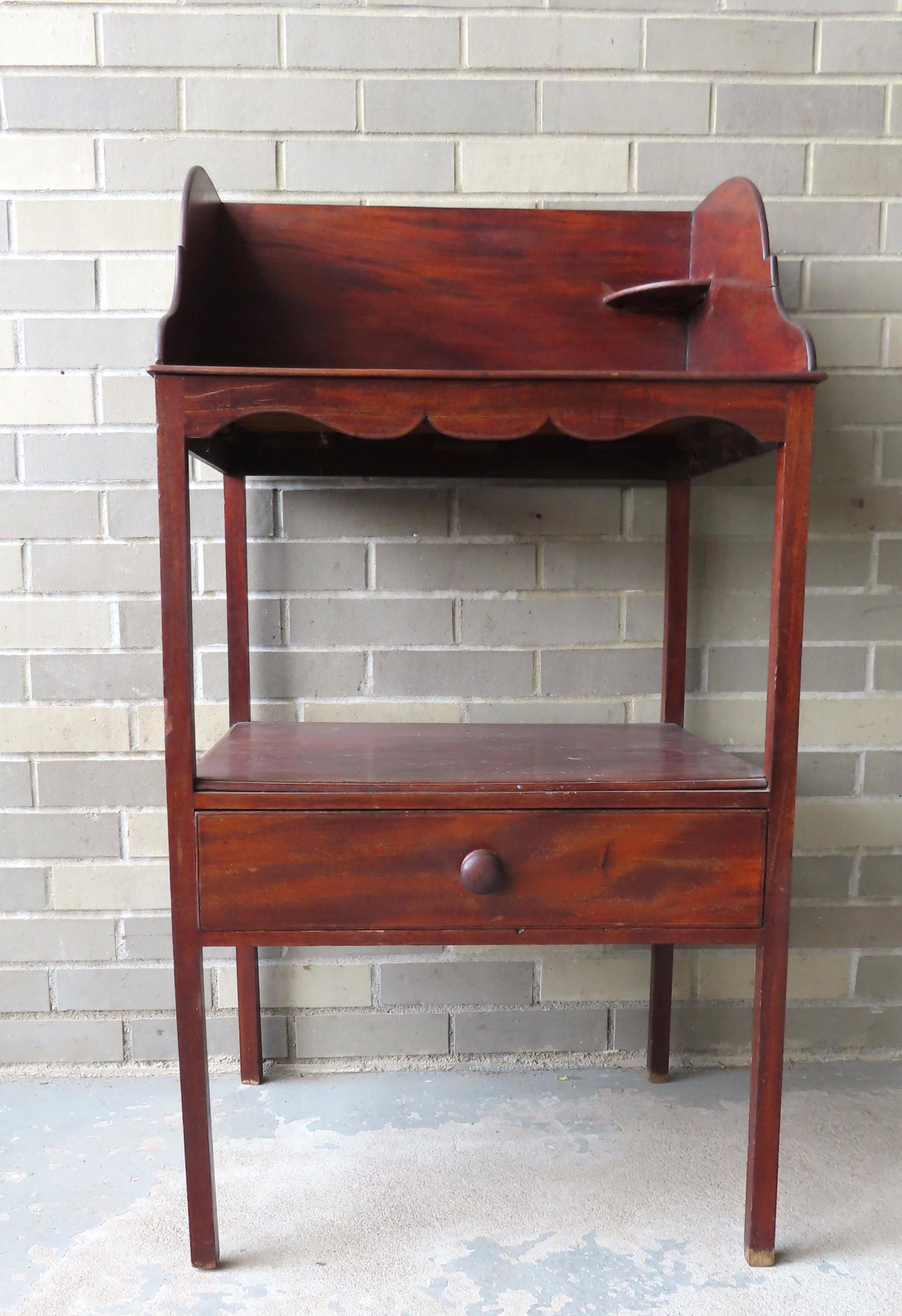 A Hepplewhite mahogany washstand with a shaped and: A Hepplewhite mahogany washstand with a shaped and dovetailed backsplash, lower shelf over drawer and having 4 narrow squared legs. Left rear leg is broken and requires professional repair, otherwise 