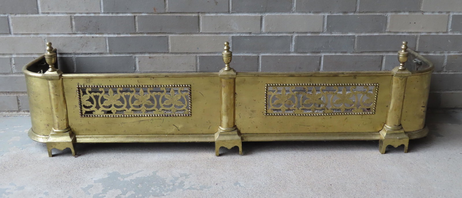 A brass fireplace fender having 3 urn finials over: A brass fireplace fender having 3 urn finials over front with openwork ornamentation of leaves on vines mounted on French feet. Measures 47 1/4"W x 13"D x 13"H. Mid to late 19th century. Very good con