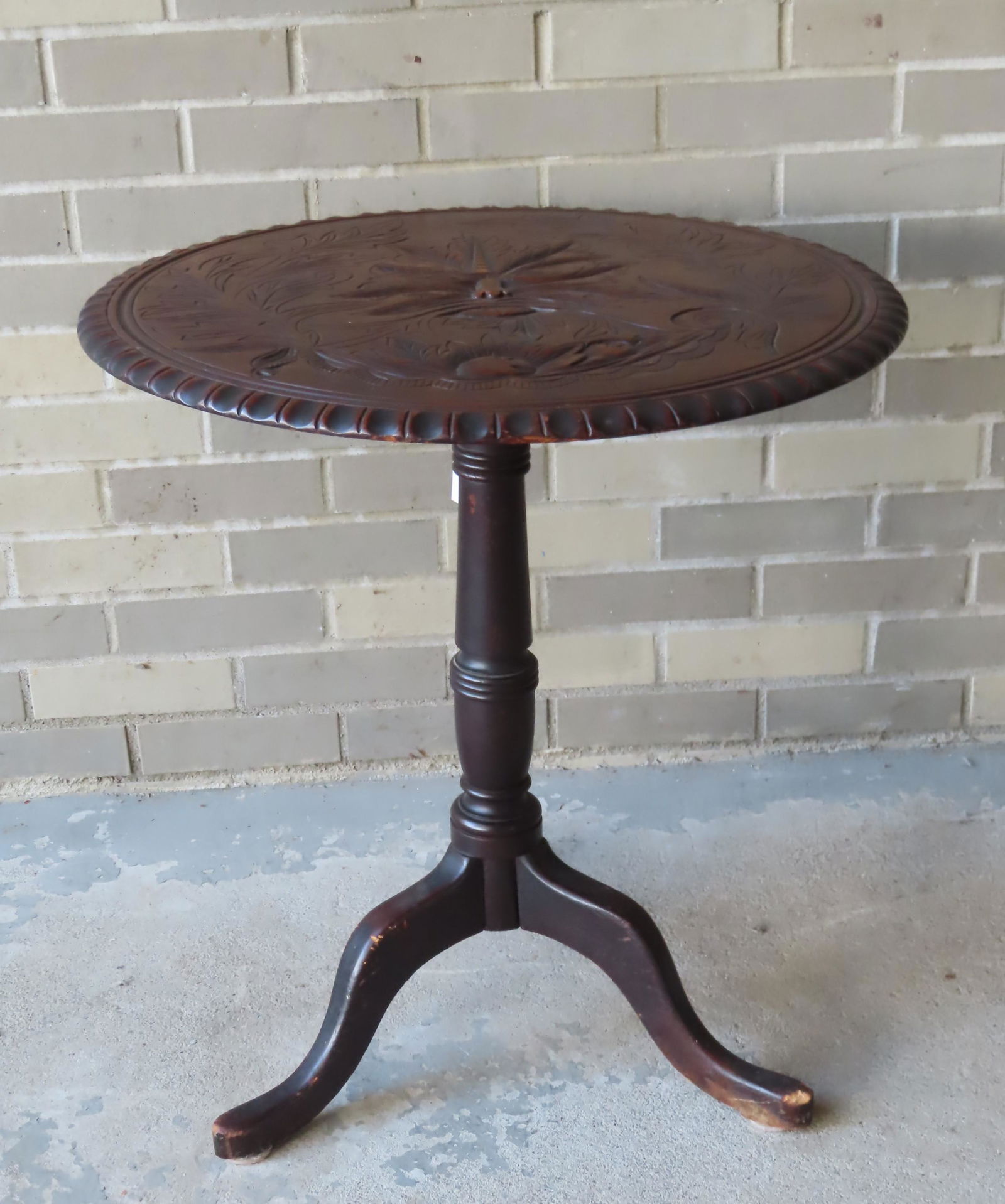Mahogany tilt-top table having a carved top decorated: Mahogany tilt-top table having a carved top decorated with a Northwind face surrounded by leaves and fruit, late 19th century. The wooden block at top of column that is hinged to the top has been repa