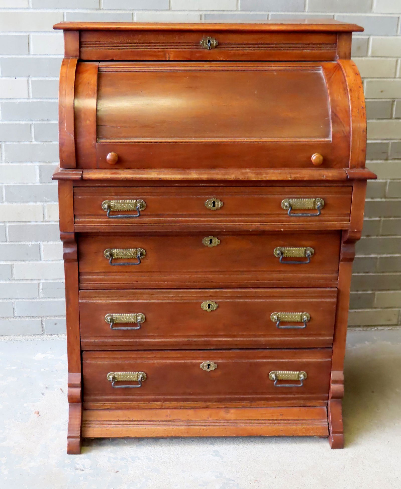 A Victorian butternut roll-front desk with fitted: A Victorian butternut roll-front desk with fitted interior with 1 drawer (pull is broken on drawer) over 4 drawers with original brass pulls, late 19th century. Condition: Locking mechanism on top wit