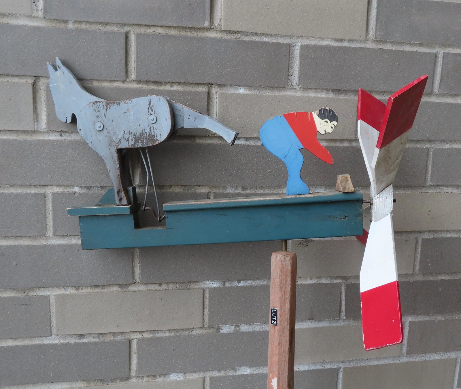 A working whirligig depicting a donkey kicking a man in: A working whirligig depicting a donkey kicking a man in the butt - the whirligig is mounted on a wooden pole with a pointed iron spear-like spike on base for insertion into the ground, mid 20th centur