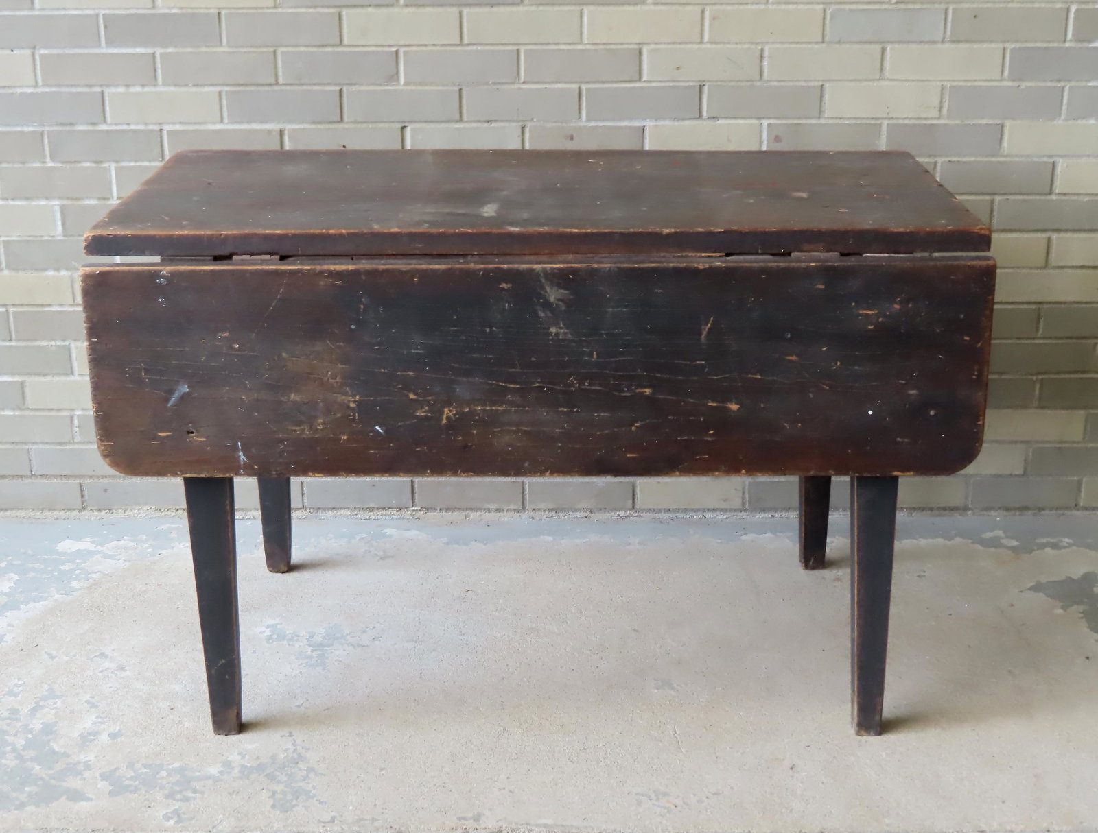 A primitive drop leaf kitchen table in old brown paint which appears to be original and having heavy: A primitive drop leaf kitchen table in old brown paint which appears to be original and having heavy tapered legs - table with paint wear but overall is very sturdy. Early 19th century. Measures 48"L 