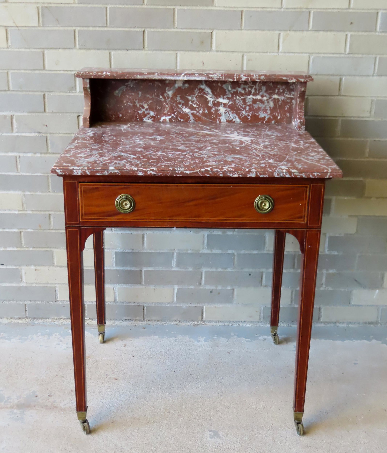 A marble top 1 drawer work table with a marble backsplash and shelf, mounted on a tapered leg base: A marble top 1 drawer work table with a marble backsplash and shelf, mounted on a tapered leg base leading to casters. Line inlays on apron, legs and beaded drawer and the marble with molded edges. Ve