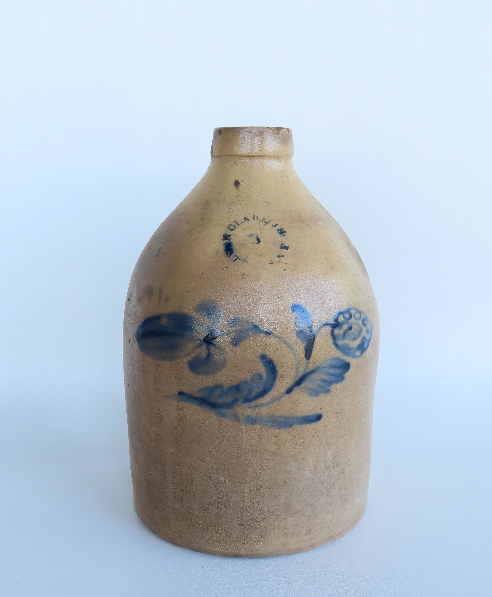 A stoneware jug decorated with a cobalt blue flower with the impressed mark of N. Clark Jr. Athens: A stoneware jug decorated with a cobalt blue flower with the impressed mark of N. Clark Jr. Athens NY, 2 gallon size. Measures 13"H x 8 1/2"W. Rim chip, pebble burst, minor staining, else good conditi