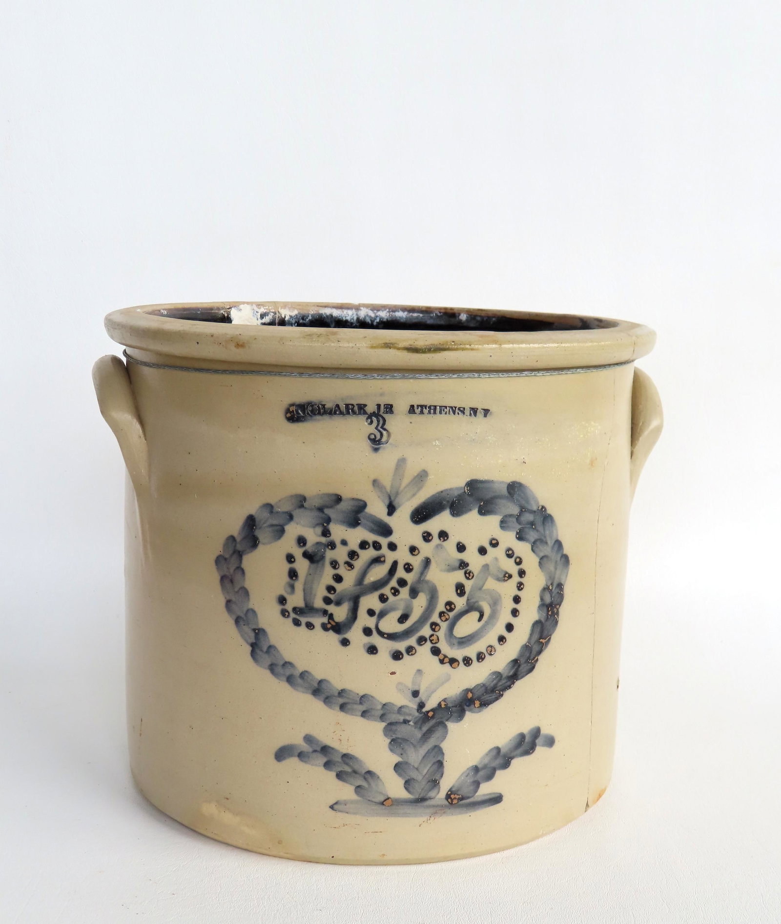 A stoneware 3 gallon crock dated in cobalt blue script 1855 within a heart shaped flower and having: A stoneware 3 gallon crock dated in cobalt blue script 1855 within a heart shaped flower and having the impressed mark of "N. Clark Jr. Athens NY". A rare example, but in poor condition having a large