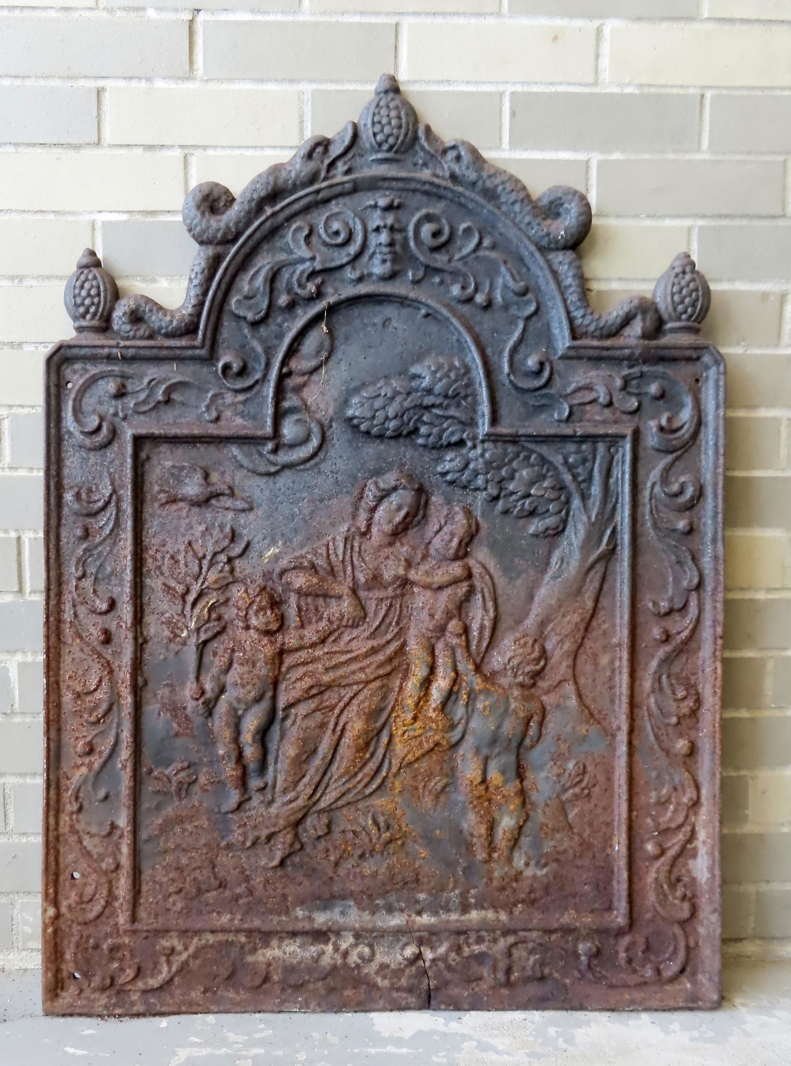 A large cast iron fire back depicting a dove above cherub offering fruit to a lady holding 2 other: A large cast iron fire back depicting a dove above cherub offering fruit to a lady holding 2 other cherubs surrounded by scrolls and a devil's face and having a domed crest decorated with serpents and