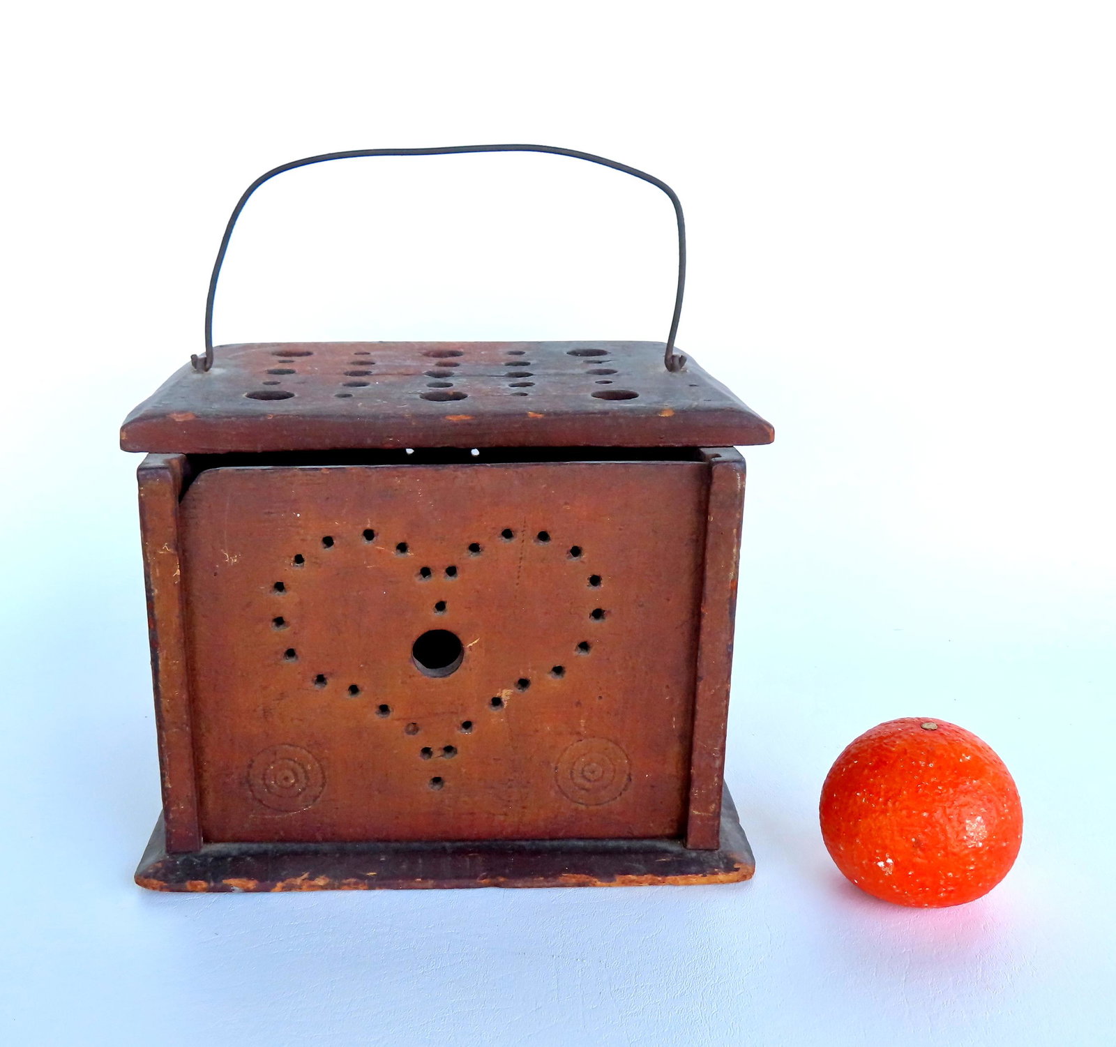 Wooden foot warmer in origingal dry red paint, with pierced heart design in removable vertical front: Wooden foot warmer in origingal dry red paint, with pierced heart design in removable vertical front door, the sides decorated with circles and having a bale handle. A large burnt hole exists in base 