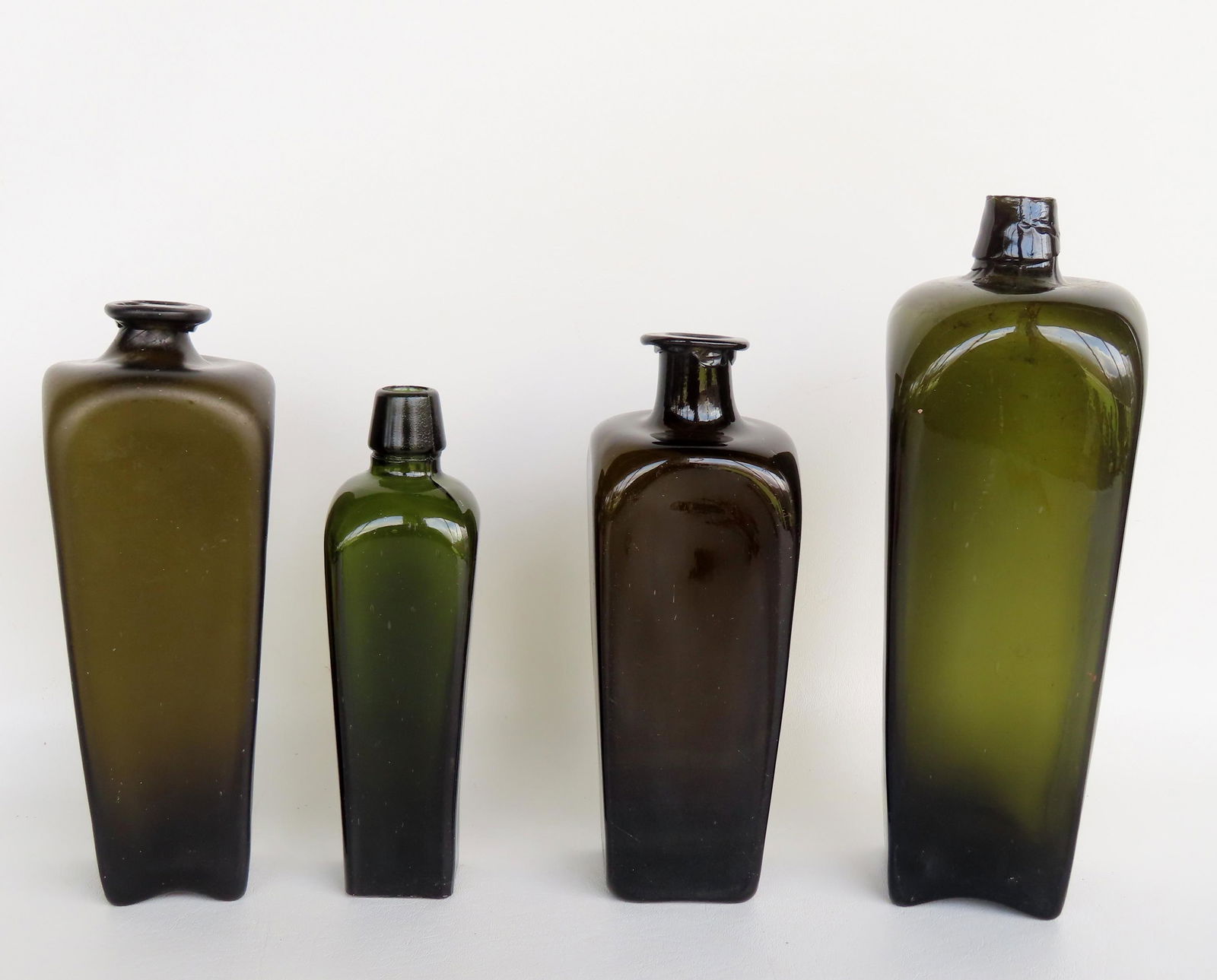Four cased gin bottles, olive green, 18th/19th century. Three with smooth pontils and 1 with scarred: Four cased gin bottles, olive green, 18th/19th century. Three with smooth pontils and 1 with scarred pontil. All in very good condition with no damage. The largest measures 11 1/4"H x 3 3/4"W.