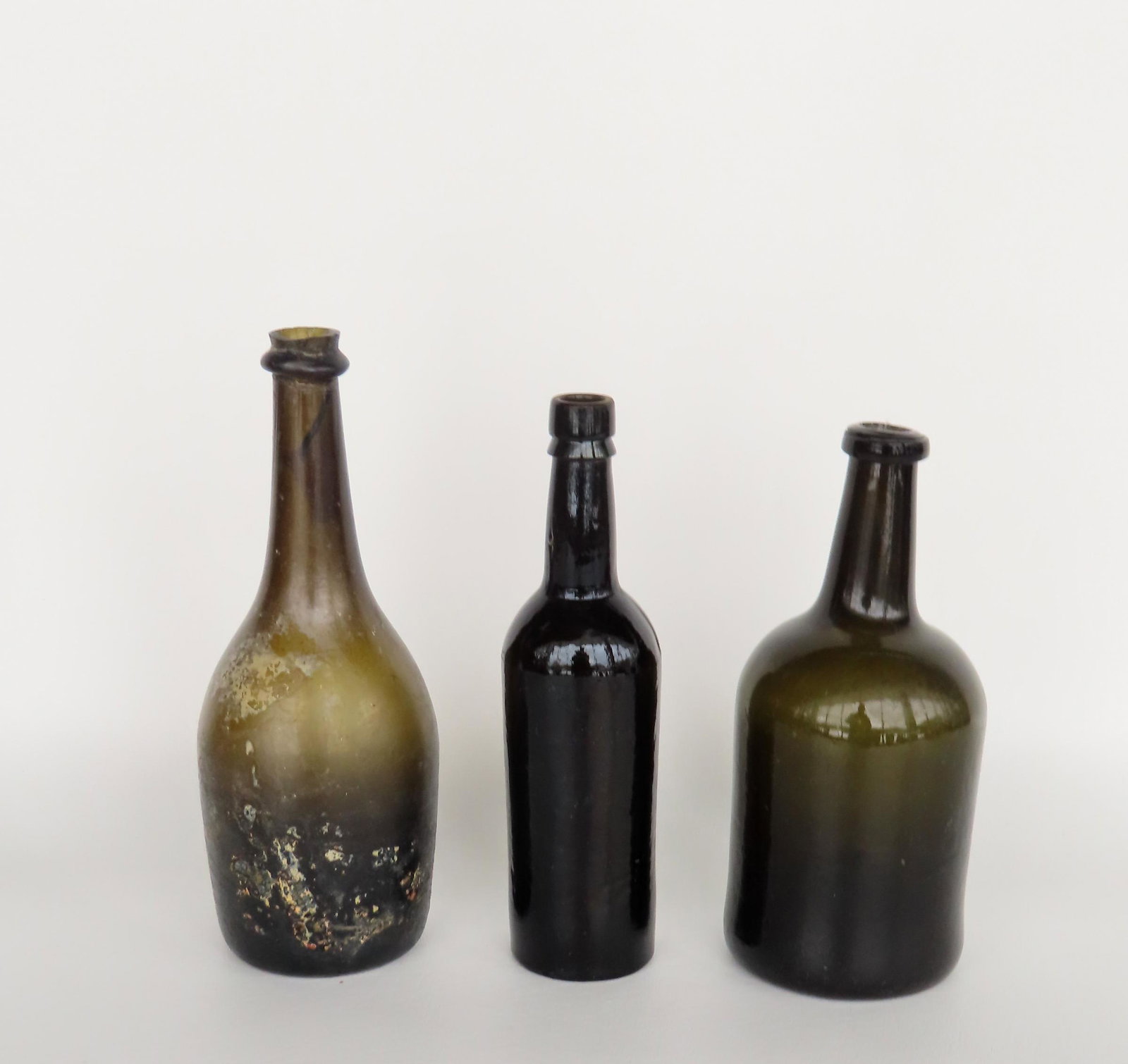 Three early wine bottles, 2 olive green and 1 dark amber, all with kicked up bottoms. The largest in: Three early wine bottles, 2 olive green and 1 dark amber, all with kicked up bottoms. The largest in irridescent olive green, scarred pontil, and with sheared mouth and string rim, 18th century - 10 1